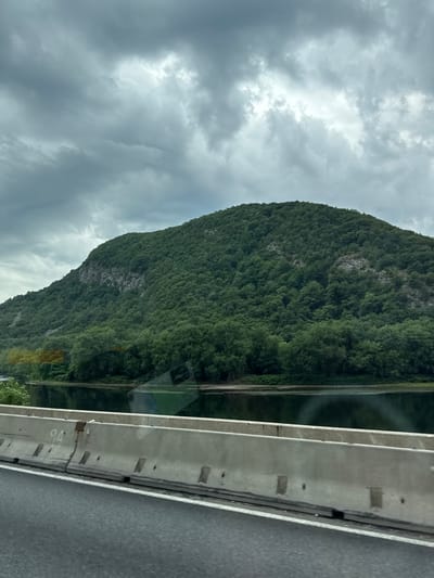 The image shows a large, tree-covered mountain rising from a body of water. The sky is overcast with dark, heavy clouds. In the foreground, a concrete barrier is visible, with a portion of an asphalt road. The scene appears to be a scenic view from a highway, likely in a natural setting. The location is identified as Hardwick Township, United States of America.
