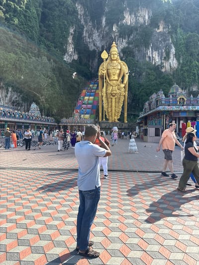The image captures a bustling outdoor scene at the Batu Caves in Selayang Municipal Council, Malaysia, under bright daylight. A colossal golden statue of Lord Murugan dominates the right side of the frame, extending from the bottom upwards. The deity stands majestically, holding a golden Vel (lance).

Behind the statue and across the left side of the image rises a massive, rugged limestone cliff face, densely covered in green vegetation. At its base, and further to the left, are vibrant and intricately decorated Hindu temple structures, showcasing a rich palette of colors and detailed carvings.

The foreground and mid-ground are populated by numerous visitors, walking on a distinctive plaza paved with interlocking geometric tiles in red-orange, grey, and light grey, creating an optical illusion of 3D cubes. In the immediate foreground, a man in a light blue polka dot shirt and jeans stands facing away, hands clasped behind his head, observing the surroundings. Further back, a man in an orange shirt and shorts, and a woman in a black top and green pants, are seen looking upwards, likely towards the statue or the caves. Many other individuals are scattered across the wide plaza, some interacting with pigeons that are present on the ground and in flight.

The clear, strong shadows indicate it is a sunny day, likely mid-morning or early afternoon. The scene captures the lively atmosphere of a significant cultural and religious site, with people engaged in sightseeing and devotion. A partial sign displaying "NATU" is faintly visible near the temple structures.