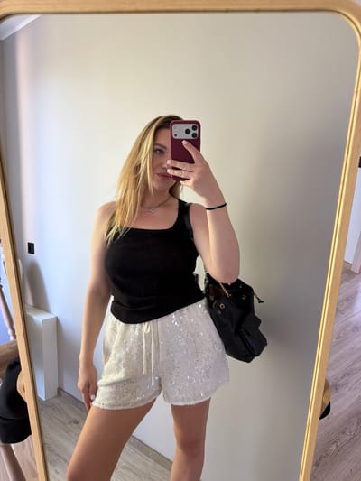 A young woman with blonde hair is taking a mirror selfie in an indoor setting. She is wearing a black tank top and white sequined shorts, with a black purse slung over her shoulder. The mirror reflects her and a portion of the room, which appears to be a dressing area or bedroom. The background is a plain wall. The lighting suggests it is daytime. The location is identified as Dreiliņi, Latvia.