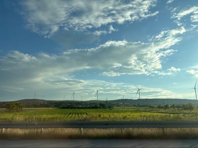 The image shows a wide shot of a rural landscape in Urbnisi, Georgia, under a bright, partly cloudy sky. In the foreground, a metal guardrail runs horizontally across the frame, separating a paved surface from a grassy shoulder. Beyond the guardrail, a field of green crops, possibly corn, stretches into the midground. In the distance, several wind turbines are visible on rolling hills. The landscape is dotted with trees and some scattered buildings. The overall impression is one of a serene, rural environment with renewable energy infrastructure. There are no people or animals clearly visible in the image.