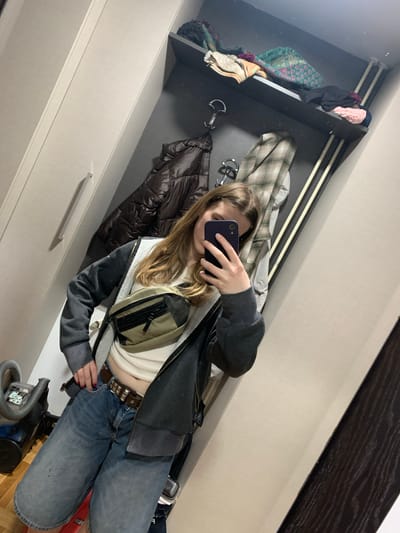 A person is captured in a mirror selfie, standing indoors in what appears to be a bedroom or dressing area in Vozdovac Urban Municipality, Serbia. Their face is largely obscured by a dark-colored smartphone held up to take the picture.

The person is wearing a casual outfit consisting of a light-colored crop top, blue denim jeans, and a partially unzipped dark gray hoodie with a lighter, possibly fleece, lining. A brown belt adorned with metal studs is visible around their waist. A light green or tan fanny pack/chest bag is worn across their torso, over the crop top. Their long, light brown hair falls over their shoulders.

The background, reflected in the mirror, shows a dark-colored shelving unit to the left, holding various folded fabrics, including a pink item and a patterned green and orange textile. Below the shelves, several items of clothing, including a brown puffer jacket and a plaid shirt, hang from hooks. To the right and bottom, the reflection reveals light-colored wardrobe or cabinet doors with silver handles. A vacuum cleaner is partially visible on the floor in the bottom right corner of the image. The overall impression is an indoor, private setting. No discernible text is visible.