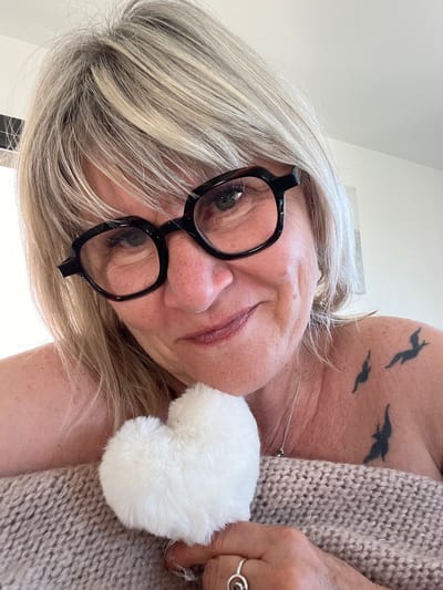 A close-up, tilted shot features a woman with short, blonde/gray hair wearing black-rimmed glasses. The subject is looking directly into the camera with a slight smile. A white, fluffy heart-shaped object is visible near her face, and she is wearing a light brown, knitted garment. On her upper chest/shoulder area, there is a tattoo depicting three birds in flight. The location is not discernable from the image. There is no way to confirm the location is Chambœuf, France.