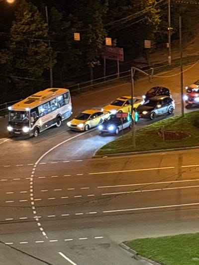 This is an aerial, nighttime photograph taken in Pushkino, Russia. The scene depicts a traffic intersection on a paved road with white lane markings. Several vehicles are visible, including a yellow bus and at least two cars. The bus appears to be a passenger bus with interior lights illuminating the cabin, suggesting people are inside. The vehicles are stopped, indicating they are likely waiting at a traffic signal. The overall lighting suggests it is dark, likely late evening or night. Some of the vehicles have their headlights on, and the reflections on the wet road surface may indicate recent rain or a damp environment. Trees and some foliage are visible on the left side of the image, bordering the road. The presence of traffic signals and multiple vehicles on a road points to an urban or suburban setting. No visible text is present in the image.