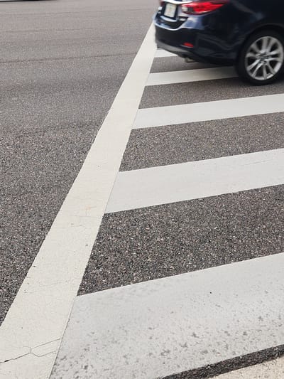 A dark-colored car is partially visible on the left side of the image, with its rear bumper and wheels showing. The car appears to be on a road or a parking lot, as indicated by the asphalt surface with white painted stripes. The stripes are typical of a crosswalk or a parking space. The image is taken from a high angle, looking down at the scene. The lighting suggests it is daytime, and the weather appears to be clear. There is no visible text in the image. The scene is static, with no discernible activity taking place beyond the presence of the car. The focus is on the textures of the asphalt and the painted lines, with the car serving as a secondary element. The overall impression is a mundane urban or suburban environment.