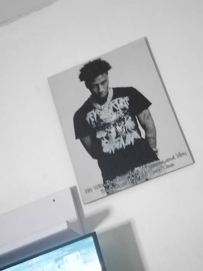 A black and white photograph is displayed on a wall, depicting a man wearing a t-shirt and a necklace. The man is positioned in the center of the photograph, with his body angled slightly to the right. He has dark, curly hair and is looking towards the camera. The t-shirt has a graphic design and text, and the necklace appears to be a chain with a pendant. Below the man's image, there is text printed on the photograph, which is partially obscured by the angle of the display. The photograph is mounted on a lighter-colored surface. The wall appears to be white. To the left of the photograph, a white shelf-like structure is visible, and below that, a portion of a television screen with a bright, light-blue hue is also visible. The scene suggests an indoor setting, possibly a room in a home or a studio. There is no information in the image to indicate a specific location in Du, Nigeria.