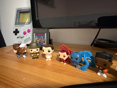 In Sankt Augustin, Germany, a collection of Stranger Things Funko Pop figures are arranged on a wooden surface. From left to right, a figure in a sailor outfit is visible, followed by one wearing a brown hat and jacket. Next is a figure in a white dress, then a figure in a red outfit with long, flowing hair. A blue, creature-like figure is positioned behind the red-haired figure, and a figure holding a basketball in white athletic wear is at the far right. Behind the figures, a grey Nintendo Game Boy and a Super Mario Land game cartridge are placed against a white wall. The scene appears to be indoors, possibly on a desk or shelf, with a dark monitor in the background.