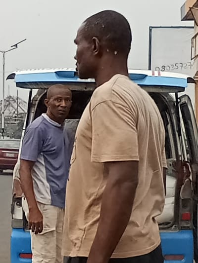 The image displays an outdoor street scene in Owerri, Nigeria, under an overcast sky. Two adult males are present near a parked commercial vehicle.

In the foreground, an adult male with dark skin and a short haircut is shown from the back-right, in profile, facing towards the right side of the frame. He wears a plain tan short-sleeved t-shirt. Directly behind him, slightly to the left, a second adult male with dark skin and a short haircut stands, looking directly at the camera with a neutral expression. He is wearing a blue and white two-toned short-sleeved shirt and light-colored trousers.

Behind the two men is a white commercial van or minibus, distinguished by a blue trim on its roof rack/spoiler area and a blue rear bumper. The rear passenger door on the vehicle's right side is open. In the background, to the left, a street light pole is visible. Further left, a portion of a reddish vehicle is present. To the right of the commercial van, a white rectangular sign or board displays a partially visible Nigerian phone number, "08035727." The ground appears to be paved.
