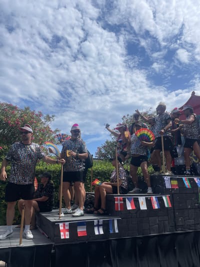 A group of people are on a float in what appears to be a Pride parade in Sitges, Spain. The float is decorated with flags and the people are wearing colorful, sparkly outfits. Some of the individuals are holding rainbow fans and drinks. The sky is blue with white clouds. The float has a stone-like texture and is decorated with various flags. Some individuals are standing and waving, while others are sitting.