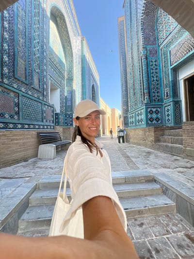 A woman wearing a cream-colored baseball cap and a white linen shirt is taking a selfie while walking through a courtyard lined with intricately tiled blue buildings. She has dark hair and is smiling at the camera. She is carrying a cream-colored tote bag. The buildings are decorated with geometric patterns and calligraphy in shades of blue, green, and white. Stone pathways and steps lead through the courtyard. In the background, other people can be seen walking. The architecture suggests this is the Shah-i-Zinda necropolis in Samarkand, Uzbekistan, known for its stunning tilework. The sky is a clear blue.