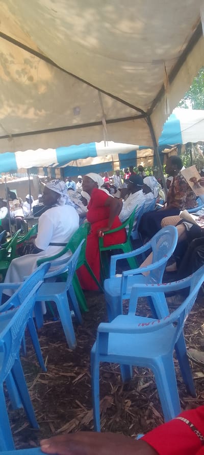 The image shows a gathering of people seated on blue and green plastic chairs under a white tent. The tent is supported by metal poles and has a blue stripe on the edges. There are shadows cast by the tent’s roof onto the ground. The ground is covered with brown leaves. There are people in the background but they are obscured by the chairs and the tent. The image was taken in Lubao, Kenya, at coordinates 0.384 latitude and 34.797 longitude.  A woman wearing a white shirt and a white head covering sits on a green chair in the center of the image. She has a white sash around her waist and a white handbag hanging from her shoulder. She is seated to the right of another woman wearing a red dress and a white head covering. This woman is seated on a blue chair and has a green chair to her right. The woman in the red dress is facing forward. The woman in white is looking at the camera. The woman in white is wearing a black belt and is sitting with her legs crossed. The woman in the red dress is sitting with her arms crossed. There are additional people in the background, but they are not clear. There is a  white plastic water bottle on the ground next to the woman in the red dress. There is a blue chair in the foreground. The camera is close to the ground and pointed upward towards the people.  The image was taken in a field or outdoor setting, with some trees or vegetation visible in the background. The  lighting is bright, suggesting a sunny day.