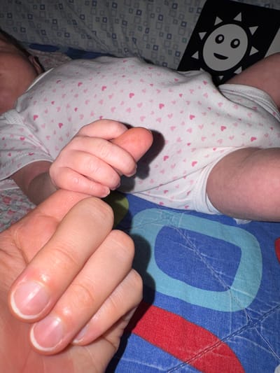 A baby, wearing a white onesie with pink heart patterns, is lying on a bed with patterned sheets. The baby is holding the pinky finger of an adult's hand. Beside the baby is a black and white fabric book with a smiling sun design. The precise location within Aprilia, Italy, cannot be determined from the image.