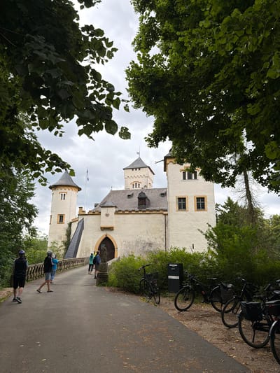 The image shows a view of a castle in Heiligenstadt in. OFr., Germany. In the foreground, a paved path leads to a stone bridge with a decorative railing. Several people, dressed in casual attire and some wearing helmets, are walking across the bridge towards the castle. To the right of the path, a row of parked bicycles is visible. The castle itself is a light-colored building with multiple towers and arched entrances. It is surrounded by lush green trees, with the leaves of some trees hanging into the frame from the top. The sky is overcast.