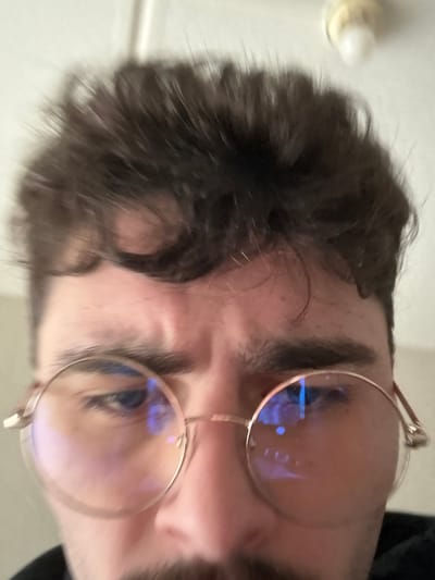 An extreme close-up, low-angle shot features the upper face of a male individual, likely a young adult. The subject has dark, curly hair falling over his forehead, thick dark eyebrows, and visible horizontal creases on his brow, suggesting a furrowed expression. He wears round, gold-framed glasses; the lenses exhibit distinct blue light reflections, possibly from a digital screen or artificial light source. His eyes appear dark behind the lenses, and the bridge of his nose is visible along with a dark mustache. The background is uniformly light-colored and out of focus, revealing a portion of a ceiling with a blurry, cream-colored fixture, possibly a light bulb socket, in the top right. The environment is an indoor setting, but the specific location within the city of Rennes, France, cannot be determined from the visual information provided. No direct interactions are depicted.