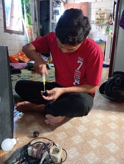 A male individual, dressed in a red short-sleeved t-shirt with white text and black long pants, is seated cross-legged and barefoot on a light brown, geometrically patterned tiled floor. He is actively engaged in repairing a small, black electrical component, holding it in his left hand while using a yellow-handled screwdriver in his right hand to manipulate its internal mechanism. His attention is focused downward on the task. To his left on the floor are a white light bulb, a tangled bundle of electrical wires, a beige multi-socket power strip, and a black plastic bag containing additional items. A large, dark rectangular object is partially visible at the extreme left. The background features a window with wooden framing and green and white patterned curtains on the left, an unmade bed or pile of textiles behind the individual, and a rear wall with white rectangular tiles and a shelving unit displaying various bottles and containers. A dark object, possibly a helmet, rests on the floor to the individual's right. The scene is illuminated by natural light entering from the left. This event occurs in Baron, Indonesia.
