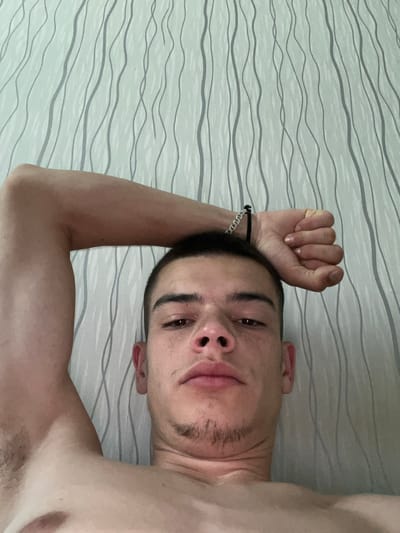 A young man, appearing to be in his late teens or early twenties, is captured in a selfie-style, close-up shot. He has short, dark hair and fair skin, with some visible stubble on his chin. His left arm is raised and bent, with his fist clenched and resting on his forehead. A silver chain bracelet is visible on his left wrist. The background consists of a wall with a light-colored, textured wallpaper featuring vertical, wavy grey lines. The lighting suggests it might be daytime, possibly indoors. There is no discernible information within the image to indicate a specific location in the city of Sopot, Bulgaria.