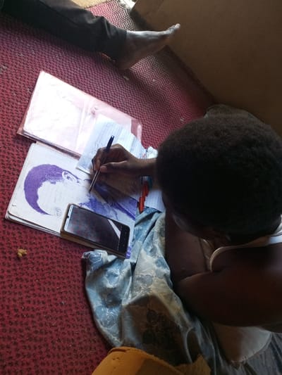 A high-angle shot captures an individual, seen from behind, sitting on a red, textured floor covering, likely a mat or carpet. The person, who appears to be a child or young adult with dark, curly hair, is focused on writing or drawing on a piece of paper with a blue pen. They wear a light-colored top and are partially covered by a light blue fabric with a floral or patterned design.

On the floor in front of them are several items. A notebook displays a prominent purple print or drawing of a person's profile. A black smartphone with a visibly cracked screen rests on top of some papers. Two clear plastic folders are also present, slightly above the main cluster of papers. In the upper left portion of the image, a bare foot and a leg covered in dark pants are visible, indicating another person is nearby.

The scene takes place indoors, possibly in a home in Aramoko-Ekiti, Nigeria, under natural daylight. A small, irregular brown object lies on the red floor in the lower left, while a larger light brown object, possibly part of a cushion or furniture, is visible in the bottom right. The primary activity depicted is the individual's engagement in a creative or academic task. No specific text is clearly legible on the papers, only patterns and marks.