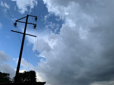 This image captures a utility pole silhouetted against a dramatic sky. The pole, a dark vertical element on the left side of the frame, supports multiple wires and insulators. It appears to be a power or telephone pole. In the foreground, at the very bottom, a dark, corrugated roofline and the dark silhouettes of trees are visible, suggesting a built-up area, possibly a residential or semi-rural setting.
The sky is the dominant feature, filled with a mix of blue sky and heavy, dark clouds, indicating dynamic weather conditions, perhaps approaching rain or a storm. Patches of brighter blue peek through in the upper left, contrasting with the ominous, gray masses that cover a large portion of the frame. The lighting suggests it is likely daytime, but the presence of dark clouds creates a moody atmosphere. There are no visible people or text in the image. The scene evokes a sense of anticipation, perhaps due to the impending weather. The location context of Kuru Karama, Nigeria, places the image in a specific geographic setting, but no direct visual cues within the image itself confirm this.