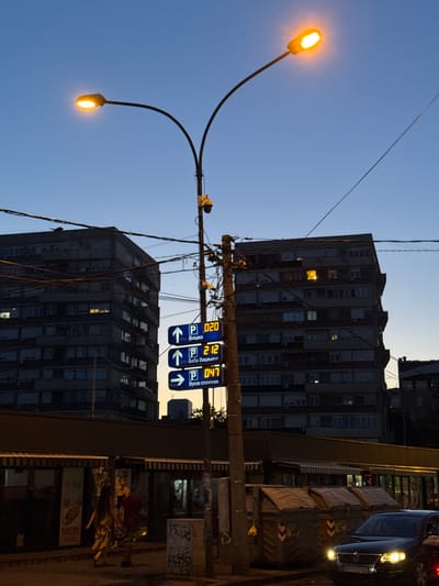 This image captures an urban street scene at dusk in the Vracar Urban Municipality, Belgrade, Serbia. The sky transitions from a deep blue overhead to a lighter, warm orange-yellow near the horizon line. Artificial lighting is prominent, including streetlights, illuminated windows in buildings, and vehicle headlights.
A central, tall utility pole dominates the midground, laden with numerous tangled black wires and equipped with at least two surveillance cameras, one yellow and one dark grey. Attached to this pole are three vertical digital parking signs displaying real-time parking availability and directional information in Cyrillic. The top sign, with an upward arrow, shows "P 020 Вишка" (P 020 Viška). The middle sign, also with an upward arrow, indicates "P 212 Баба Вишњина" (P 212 Baba Višnjina). The bottom sign, with a rightward arrow, displays "P 047 Вуков споменик" (P 047 Vukov spomenik). The names "Baba Višnjina" and "Vukov spomenik" confirm the location within Vracar.
Two large, multi-story buildings, likely residential or mixed-use, form the background. The building on the left is prominent, featuring numerous windows, some with warm interior lighting and visible air conditioning units. Below these buildings, at street level, are single-story storefronts with awnings, one of which has a visible "BAKERY" sign.
On the sidewalk in the lower left, two individuals are walking. One person with long hair, wearing a light-colored garment, is moving from left to right.
In the lower right foreground, a dark-colored car with illuminated headlights faces the viewer. To its right, a portion of a red vehicle is visible. Three large, rectangular, dark-colored industrial waste bins with orange and white markings are situated on the sidewalk behind the car. One bin has a yellow plastic bag hanging from it. The base of the utility pole exhibits graffiti. A dual-arm streetlight with two glowing yellow-orange lamps curves from the central pole in the upper left, providing street illumination.
