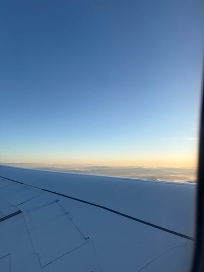 The photograph was taken from inside an airplane, looking out of a window and down at the ground. The lower right of the frame is filled by a portion of the airplane's wing, which appears to be white or light gray in color. Visible are panel lines and small rivets on the wing's surface. The sky is visible on the left side of the frame, transitioning from blue to a lighter yellow near the horizon. Along the horizon, there is a layer of haze or thin clouds giving a soft appearance to the area where the sky meets the earth. The location is possibly above Moreira, Portugal, which cannot be fully determined by the image provided.
