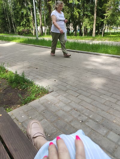 In Navapolatsk, Belarus, an elderly woman in a white t-shirt and green pants walks along a paved park path, carrying a white plastic bag. She is walking towards the left side of the frame. To the left of the path, a park bench is visible, occupied by a person whose legs and feet are in the foreground, wearing a light-colored shoe and a light blue garment. Their fingernails are painted a bright pink. Behind the path, a grassy area and a line of trees form the background. Sunlight dapples the paved walkway.