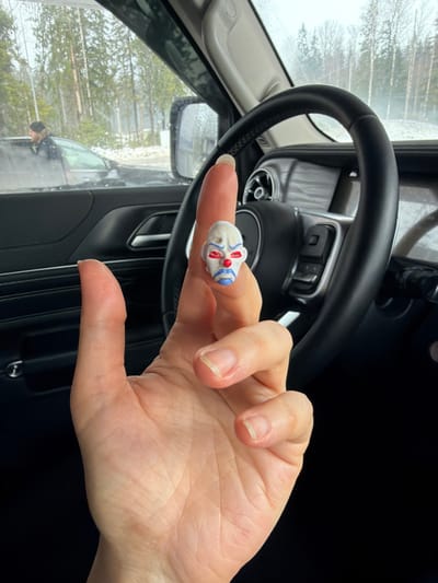 A forensic examination of the image reveals a close-up perspective from the interior of a vehicle, displaying a human hand holding a small, white miniature head, against a backdrop of an outdoor scene in Kissolovo, Russia.

**Scene Description:**
The foreground is dominated by an open, light-skinned human hand, palm facing outward, with fingers slightly curled. A small, white figurine head, approximately 2-3 cm tall, is balanced on the tip of the extended index finger. This miniature head depicts a stylized clown-like face with red markings above the eyes, red eye sockets, a red circular nose, and blue markings below the eyes extending to the chin, resembling the Joker character's mask from "The Dark Knight." The fingernails of the hand appear natural and unpolished.

**Interior Environment:**
The hand is positioned in front of a dark-colored vehicle interior. A black, leather-wrapped steering wheel, equipped with controls, is visible on the right side of the frame, partially obscured by the hand. The vehicle's dark dashboard and door panel are also visible, suggesting the image was captured from the driver's or front passenger's seat.

**Exterior Environment (Kissolovo, Russia):**
Through the vehicle's windshield and side mirror, a wintry or early spring outdoor environment is visible. Tall, bare deciduous trees and conifers, indicative of a forest or wooded area, line the background. Patches of snow are present on the ground. A paved road or driveway runs horizontally across the mid-ground. To the left, a dark-colored sedan or SUV is partially visible, with its driver's side window down. A male individual with dark, possibly curly hair and a beard, wearing a dark jacket, is observed leaning into or looking toward the driver's side of this external vehicle.

**Actions/Interactions:**
The primary action is a hand presenting a miniature object. The individual outside the other vehicle appears to be observing or interacting with the vehicle.