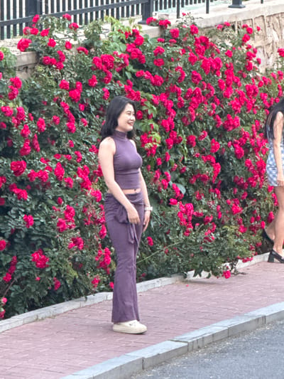 A medium-angle shot of a woman in a purple outfit standing on a brick sidewalk. The background features a wall of red flowers. A portion of a metal fence is visible on the left side. A partial view of another person's legs appears at the top of the image. The location is Zhongtun, China.