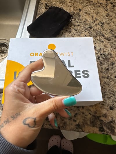 A close-up, high-angle photograph shows a human hand, adorned with vibrant teal nail polish, holding a reflective, silver-toned, heart-shaped gua sha tool. The back of the hand displays two tattoos: a black outline of a heart and partially visible script that reads "THE SUN" (reversed). A thin gold-colored ring is visible on one of the fingers.
The gua sha tool is held over a white product box featuring an orange circular logo and text. Visible portions of the text include "ORANGE TWIST" on the side and larger letters "AL ES" on the top surface. A black drawstring pouch lies to the left of the box.
The objects rest on a speckled, light brown granite-like countertop. A stainless steel kitchen sink with a drain is visible in the lower left quadrant of the frame. In the background, a pair of light pink clogs and a green object are partially visible. The scene is an indoor setting, likely a kitchen or utility area, under bright, diffused lighting. The photo was taken in Seal Beach, United States of America.