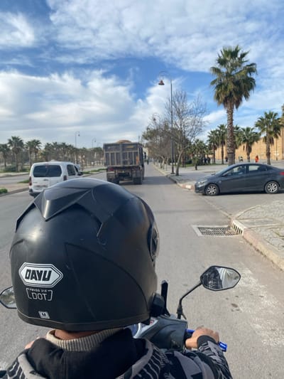 The image depicts a first-person perspective from a person riding a scooter or motorcycle on a paved road in Mechouar Fes Jdid, Morocco, under a partially cloudy blue sky.
In the immediate foreground, the back of a black motorcycle helmet is visible, featuring white "DAYU", "FMVSS 218", "DOT", and "M" (size) labels. Below the helmet, the upper back of the rider, wearing a grey and black patterned hoodie over a light-colored turtleneck, is present. The rider's hands are visible on the handlebars, with a blue grip on the right side. A rearview mirror on the right reflects a distorted image, appearing to show a white and black object or person.
Ahead on the asphalt road, a white commercial van, possibly a Peugeot Partner, is visible in the left lane. Further down the same lane, a large dark-colored dump truck, carrying a load of light-colored material (likely sand or dirt), proceeds away from the camera. On the right side of the road, a dark grey sedan, resembling a Hyundai Accent, is parked or stopped with its front wheels slightly angled to the left. A concrete curb and a metal storm drain grate are visible on the right shoulder.
The environment is characterized by numerous tall palm trees lining both sides of the road, forming rows that extend into the distance. Bare deciduous trees are also interspersed among the palm trees on the right. Ornate streetlights with curled metal designs are spaced along the road. In the far background on the right, a substantial, light-brown colored historic wall, indicative of a fortification or city rampart, is visible behind the palm trees. Several indistinct human figures are discernible near this wall. The overall scene suggests a main thoroughfare within an urban or semi-urban area.