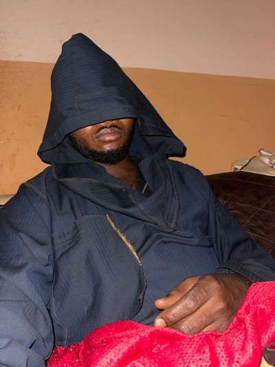 A dark-skinned individual lies reclined, largely covered by a dark blue hooded robe or garment. Only a portion of their face, including the mouth and beard, is visible beneath the hood. A hand, also dark-skinned, rests on a red, textured blanket or piece of fabric. The person is positioned against a light tan wall and what appears to be a brown leather couch. The image is rotated, so the person is laying horizontally.