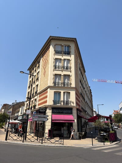 An exterior, low-angle, wide shot captures a bright, sunny day in Courbevoie, France, focusing on a triangular building at an intersection. The building is several stories high, with a façade alternating between yellow brick with a red diamond pattern and cream-colored stucco. Numerous windows with black wrought-iron balconies punctuate the structure. At street level, a boulangerie with a bright pink awning is visible, displaying signage. To the left of the boulangerie, other shops with awnings and signs are present, including one with a Total Energies logo. Street signs indicate "Boulangerie Pâtisserie," "Centre Hospitalier Rives de Seine," and "Site de Courbevoie." A streetlamp stands prominently in front of the building. In the foreground, a street with a crosswalk is visible. A person walks with a stroller on the sidewalk. In the background, a red and white construction crane is visible against the clear blue sky.