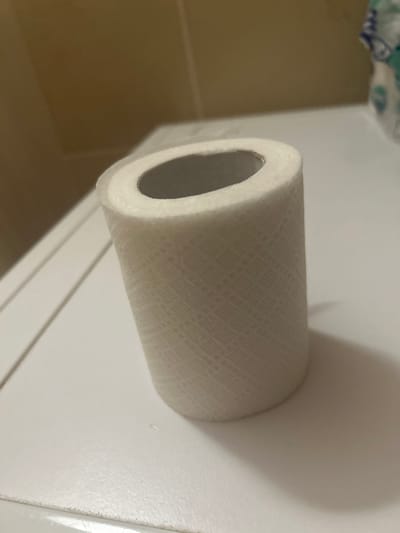 A roll of white toilet paper sits on a white, flat surface. The toilet paper has a subtle textured pattern. The background is out of focus, with a brown tiled wall visible on the left and a white surface with some blue and white packaging on the right. The scene appears to be indoors, possibly a bathroom or laundry room. There is no indication of the city of Novokuybyshevsk, Russia, or any people in the image.