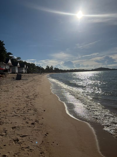 The image depicts a sun-drenched beach scene at Baan Mae Nam, Thailand. The sun is high in the sky, casting a bright glare on the water and creating sparkling reflections. Gentle waves lap onto the sandy shore, leaving a thin line of white foam. The beach is lined with palm trees and vegetation, with a few buildings visible in the background, possibly resorts or bungalows. Footprints are visible in the sand, suggesting recent activity. A surfboard stands upright on the beach near a sign that appears to be a menu or informational board. The sky is a clear blue with scattered, wispy clouds. The overall mood is serene and inviting, conveying a sense of a peaceful tropical vacation. The time of day is likely late morning or early afternoon, given the strong sunlight.