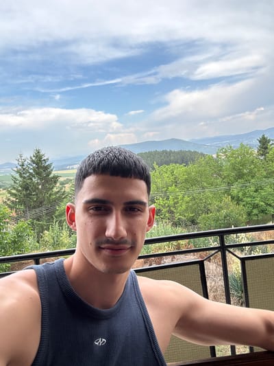 The media file shows a close-up, rotated selfie of a young man in Vladislavtsi, Bulgaria. The man has dark hair, dark eyes, and a light complexion, and is wearing a navy blue ribbed tank top. He is smiling slightly, with a faint mustache and stubble. His left arm and shoulder are visible.
The background suggests an outdoor setting with a scenic view. There are lush green trees and rolling hills or mountains in the distance. The sky is partly cloudy with wispy white clouds against a blue sky. There is a black metal railing in the foreground, suggesting the man is on a balcony or terrace. The time of day appears to be daytime, possibly late morning or afternoon, given the lighting. The weather seems pleasant and clear.
The image contains a small white logo on the tank top, but no other visible text. The overall impression is one of relaxation and enjoying a scenic view.