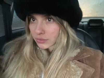 A woman with blonde hair is looking down at the camera. She is wearing a brown jacket with fur trim and a black fur hat. She appears to be indoors, possibly in a vehicle. The location is unknown, and there is no information to suggest this image was taken in San Fernando del Valle de Catamarca, Argentina.