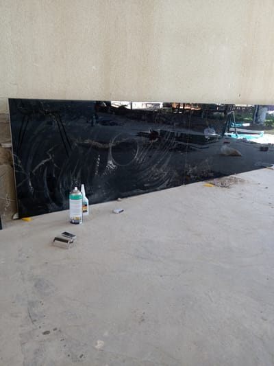 The image shows a large, black, reflective surface leaning against a textured, beige wall. The surface appears to be a slab of polished stone or glass, possibly granite or marble, with swirling patterns etched into it, likely from a cleaning or polishing process. In front of this slab, on a concrete floor, are several objects. These include a can of spray, a white plastic bottle, a small box of matches, and two small, rectangular objects with textured surfaces, possibly cleaning blocks or erasers. The concrete floor is rough and shows some signs of wear and staining.

In the reflection on the black surface, a busy outdoor scene is visible. There are glimpses of people, construction materials like bags of sand or gravel, and some blue and green equipment, possibly for construction or landscaping. The background suggests an open area, with hints of trees and structures. The lighting indicates it is daytime, with sunlight casting shadows.

The overall scene suggests an ongoing project or a work in progress, possibly related to construction, renovation, or the installation of the black surface. The presence of cleaning supplies and the polished finish of the slab imply an effort to prepare or maintain it. The location context provided, Buka, Uzbekistan, adds a geographical detail to the scene.