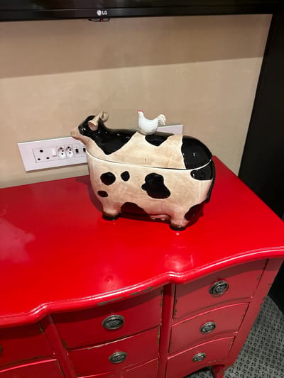 The image shows a close-up of objects in an indoor setting. A red dresser with decorative drawer pulls is prominently featured on the right side of the image. Next to the dresser, on a light-colored wall, is a ceramic cow with a chicken figurine attached to it. The cow appears to be a decorative object, possibly a piggy bank or similar item. An electrical outlet is visible below the cow. The left side of the image shows a black television set mounted on the wall. The location of the image is not discernible from the image itself, and the city of Cape Town, South Africa cannot be determined from the available information.