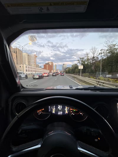 The image provides a driver's perspective from inside a vehicle, likely a Jeep Wrangler, on a multi-lane highway in Boston, Massachusetts, United States. The vehicle is traveling southbound on I-93 (Southeast Expressway), with the view extending towards the South End and the broader Boston skyline.
The time of day is late afternoon or early evening, indicated by the overcast sky with scattered clouds and diffused natural light. The vehicle's instrument cluster shows a speed of 21 mph, the transmission in 'PRND', and an odometer reading of 23,191 miles. The fuel gauge indicates approximately one-quarter of a tank.
Ahead, the highway carries moderate traffic, with several passenger vehicles, including a white Jeep Wrangler in the adjacent left lane, moving at a slow speed. Brake lights are illuminated on some vehicles in the distance.
On the left side of the highway, a concrete barrier separates the roadway from urban development. Visible structures include a large, light-colored building with numerous windows, followed by a cluster of red-brick buildings, and a modern high-rise tower further in the distance, contributing to the city skyline. A streetlight is visible above the left lanes.
On the right side of the highway, another concrete barrier runs parallel to the road, separating it from a lower-lying area. This area is densely populated with mature trees and foliage. A white rectangular sign with black text reading "EMERGENCY STOPPING ONLY" is mounted on a pole within this treed section. Partially visible through the trees are elements consistent with a railway line, including tracks and signaling equipment.
The upper portion of the vehicle's windshield displays a faded, rectangular adhesive residue or sticker, and the overhead visor area contains a "WARNING / MISE EN GARDE" label.