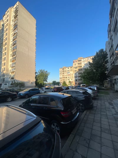 The image depicts a residential street in Yambol, Bulgaria, confirmed by the "Y" regional code on the license plate "Y 7755 BK" visible on a black Mercedes-Benz B-Class. The scene is dominated by two large, multi-story apartment buildings, characteristic of socialist-era architecture, under a clear, bright blue sky.

On the left, a tall, light-colored residential block with numerous windows and balconies rises, casting a shadow on its lower facade. Several cars are parked in the foreground and midground, including a dark sedan and a blue SUV.

On the right, a paved area with a distinct diamond-patterned sidewalk extends into the midground, where more vehicles are parked closely together, including a silver hatchback and a white SUV. Behind these cars, another, slightly shorter apartment complex is visible, partially obscured by a mature green tree. A white van is discernible in the distance.

The lighting suggests a morning or late afternoon sun, creating elongated shadows. No individuals are visible within the frame. The environment is urban, indicating dense residential living and on-street parking. A thin wire or distant object crosses the sky in the upper right.