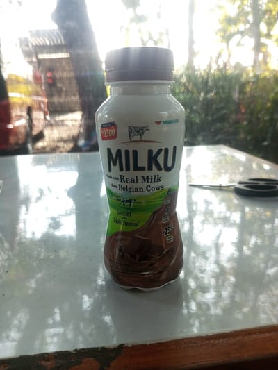 This media file showcases a bottle of "MILKU" chocolate-flavored milk, positioned on a reflective, light-colored surface that appears to be a table. The bottle's label is prominent, displaying the brand name "MILKU" in large, bold letters. Below it, text indicates it's "Made with Real Milk from Belgian Cows." Additional text on the label provides nutritional information, stating "6 Sumber Vitamin," "4 Sumber Mineral," and "25% Kalsium Harian." The price of Rp 3500 is also visible on the bottle. The background is blurred but suggests an outdoor setting with sunlight filtering through trees, creating a dappled light effect. The overall impression is that of a casual, daytime scene, possibly in a garden or outdoor seating area. The lighting indicates it's daytime, with bright sunlight, and the lush green foliage in the background suggests a tropical or sub-tropical climate. The presence of the milk bottle on a table suggests a moment of refreshment or a product display in a relaxed environment. There are no people visible in the image. The scene is set in Tangerang, Indonesia.