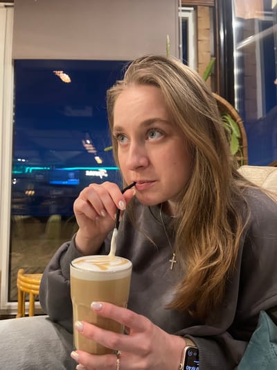 A female individual is seated indoors at what appears to be a cafe or restaurant in Yantarny, Russia, indicated by a blue neon sign with the Cyrillic word "Янтарный" reflected in the window behind her. The woman, with light-colored hair and blue eyes, is wearing a dark grey sweatshirt, light-colored pants, a gold necklace with a cross pendant, and an Apple Watch on her left wrist showing "19:40." Her fingernails are painted with a pearlescent polish, and a ring is visible on her left ring finger. She is actively drinking from a tall glass containing a layered coffee beverage with latte art and a foamy top, using a black bent straw. Her right hand holds the straw, while her left hand holds the glass. Her gaze is directed upward and to the left of the frame. The background includes a large window displaying a nighttime exterior scene with reflections of streetlights and the aforementioned neon sign, along with interior elements such as a wicker item containing green foliage and a cream-colored upholstered seat.