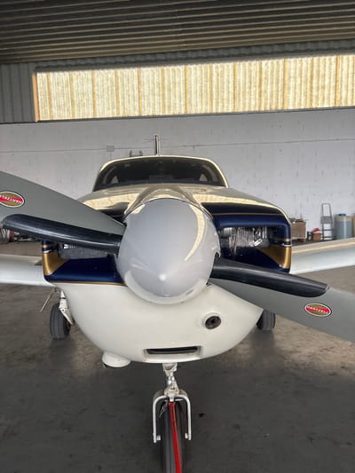 A static, head-on view of a single-engine propeller aircraft is captured inside a hangar at Toussus-le-Noble, France. The aircraft features a two-blade Hartzell propeller with a silver spinner. The aircraft's forward fuselage and cowling are primarily white, accented by a dark blue section with a gold stripe on the engine cowling. Visible engine components are partially exposed behind the propeller. The nose landing gear is extended, presenting a single wheel with a red stripe on its tire tread. The floor of the hangar is concrete, and the walls are painted white. Natural light illuminates the scene through a large translucent corrugated panel spanning the upper section of the hangar. In the background to the right, workshop equipment including a ladder, wooden crates, and plastic containers are visible. No individuals, actions, or interactions are depicted.