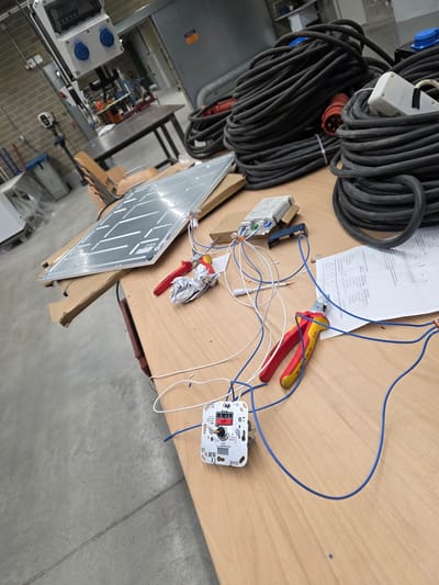 The image shows a workshop or electrical supply room, likely in Leudelange, Luxembourg, given the context of the question. A wooden workbench dominates the foreground, laden with electrical components and equipment. A large, flat, rectangular object, possibly a lighting panel, rests on cardboard boxes on the left side of the bench. To its right, a white electronic device with a red dial and blue and white wires is laid out, connected to additional wiring. A pair of red and yellow handled wire cutters rests near the electronic device, next to a crumpled piece of paper. Behind these items, large spools of thick black electrical cable are piled high. In the background, a metal cabinet and a brick wall are visible, with further electrical equipment, including a white box with blue connectors, mounted on the wall. The overall impression is of a work-in-progress or a storage area for electrical supplies.
