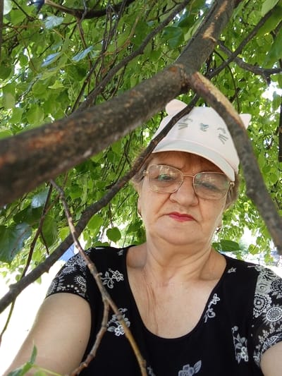 A woman with glasses and a pink cap with cat ears is looking at the camera. She is wearing a black shirt with a floral pattern. She is in a park with green trees and branches. The photo appears to be taken on a sunny day. The exact location in Ulyanovsk, Russia, cannot be determined from the image alone.