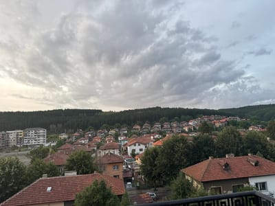 This is a high-angle shot of a residential area in Pernik, Bulgaria. The sky is overcast with gray clouds. The foreground is dominated by the red-tiled roofs of houses, some with dormer windows and chimneys. Mature green trees are interspersed among the buildings.
In the mid-ground, more houses with red-tiled roofs are visible, built on a hillside. A multi-story apartment building, gray and white with numerous windows, stands on the left side of the frame, overlooking a road. Several cars are parked on the road and in a lot adjacent to the apartment building.
The background features a dense forest of dark green pine trees covering a hill. The overall impression is of a somewhat aged, densely populated suburban or town environment nestled within a hilly, forested landscape. There are no people visible in the scene.