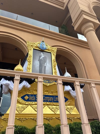 The image shows a monument with a portrait of a woman in traditional attire, placed on a gold structure adorned with black and white fabric drapes. The monument is located in front of a building with arched entrances and a light-colored facade. The text "Veda S Company Lim" is visible on the gold structure, suggesting a corporate affiliation. The scene appears to be outdoors, with green bushes visible at the bottom of the frame. The overall impression is one of a memorial or a tribute, possibly for a significant figure or event. The presence of the building's architecture and the general setting might suggest a public space within a commercial or administrative area.