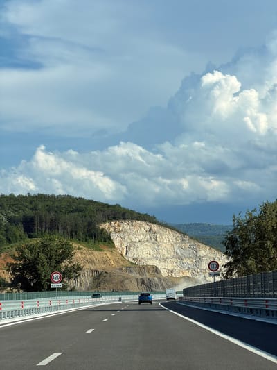 The image depicts a highway in a mountainous region, with a sheer rock face on one side and a steep, wooded slope on the other. The highway is paved with asphalt and marked with white lane dividers. A metal guardrail runs along the side of the road, equipped with reflective markers.
On the highway, a white van is visible in the distance, followed by a blue car. A speed limit sign indicating "120" is prominently displayed next to the road. Another similar sign is visible further down the road. The sky is partly cloudy, with large, white cumulus clouds against a pale blue background. The time of day appears to be daytime, likely late morning or early afternoon, given the lighting. The overall scene suggests a scenic drive through a natural landscape, possibly a quarry or a mountainous pass.
The location context provided is Peshterna, Bulgaria. The prominent rock face suggests a geological feature that might be a natural cliff or a result of quarrying activities. The dense pine forest on the slope indicates a temperate climate. The presence of the highway suggests a connection to human infrastructure facilitating travel through this terrain. The weather appears pleasant, with good visibility.