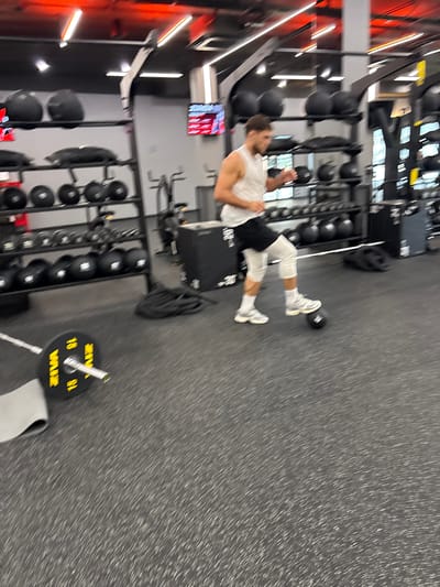 In a gym located in Chalong, Thailand, a person is performing an exercise. The person, wearing a white tank top, black shorts, and white compression sleeves and socks, is stepping on a black medicine ball. They are wearing white sneakers. In the foreground, a barbell plate labeled "15" is visible on the floor. The gym environment features rows of dumbbells, weight racks, and other gym equipment. The lighting is bright, with red accents on the ceiling. The flooring is a dark, textured material common in gyms. The image has a motion blur effect, suggesting the person is in motion.