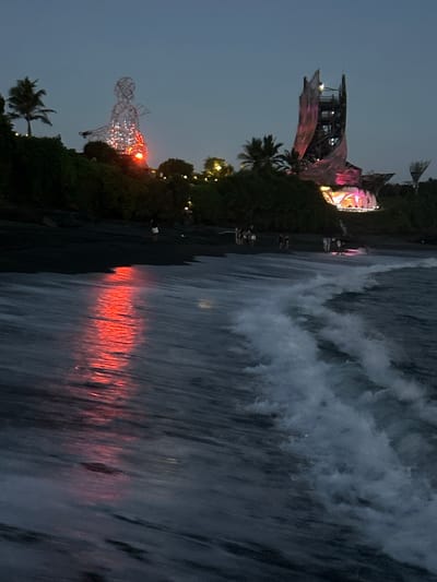The image depicts a nighttime scene on a beach in Kediri, Indonesia. The composition is divided, with a surreal, illuminated structure and lush vegetation on the left, and a turbulent ocean with crashing waves on the right. On the left side, a large, skeletal figure made of white lights stands tall, with a red light emanating from its chest. This figure appears to be part of a festival or event, as evidenced by other illuminated structures further in the background, adorned with pink and yellow lights. Palm trees and dense foliage surround the structures, suggesting a tropical environment. The sky is dark, indicating it is late evening or night.
The right side of the image is dominated by the ocean. Powerful waves are breaking onto the dark sand or shore, creating a dramatic and dynamic contrast to the static illuminated structures. The water is dark, with the white foam of the waves being the most prominent feature. A striking detail is the reflection of red and orange light on the wet sand and the surface of the water, likely from the festival lights, casting an eerie glow. The overall mood is one of spectacle and perhaps a touch of mystery, with the juxtaposition of man-made illumination and the raw power of nature. There is no visible text in the image.