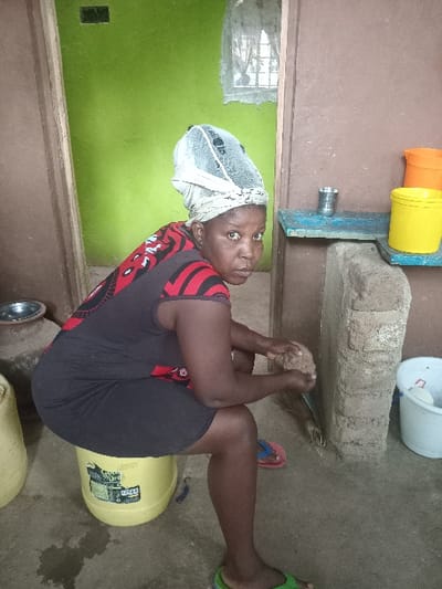 A woman is depicted in an indoor or semi-enclosed utility space, likely a kitchen or work area, in Manda-Shivanga ward, Kenya. She has dark skin and is wearing a dark, sleeveless top with red patterns, dark shorts, and a light-colored head covering. She is shown in a squatting or seated position on an upturned yellow plastic bucket, looking directly at the camera with a neutral expression.
With both hands, she is holding a small, rough, irregularly shaped object, possibly a coconut, appearing to be in the process of grating or preparing it. Near her bare feet, red and green flip-flops are visible on the concrete floor. A silver bowl and another yellow container are also on the floor nearby.
The walls are painted in two tones: an upper brown/beige and a lower vibrant green. A window on the green wall provides natural light, indicating it is daytime. Above the window line, on a raised counter-like structure made of concrete blocks and a wooden plank, are orange and yellow plastic buckets and a metallic pipe or spigot. A small broom rests on the floor near this structure. The floor is concrete. Partially visible text on the yellow bucket she is sitting on includes "GIANT GR..." and "PROD...".