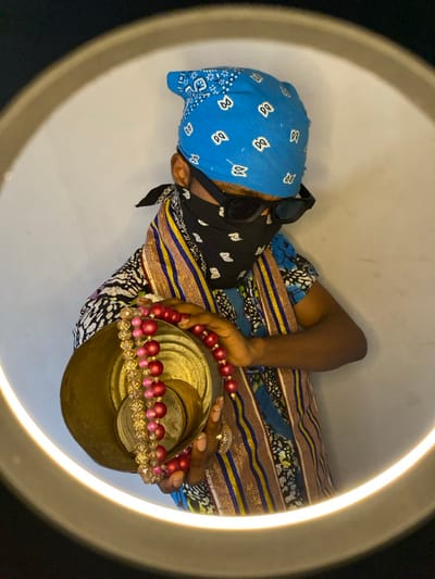 A person with obscured facial features stands centrally within a bright circular light ring against a plain, light-colored background in Ngleshi Amanfro, Ghana. The individual wears a blue bandana with white paisley patterns on the head, black framed sunglasses, and a black bandana with white paisley patterns covering the nose and mouth. A multi-colored patterned shirt, predominantly blue and white with black details, is visible, along with a striped sash or stole featuring gold, blue, and black bands. Both hands are holding a stack of at least two nested, gold-toned metallic bowls, the largest being outermost, with visible concentric texture inside. Two strands of beads are draped over the rim of the largest bowl: one strand consists of spherical, matte red beads, and the other consists of faceted, clear/pinkish-gold beads. A silver-toned ring with an ornate design is visible on the ring finger of the individual's right hand. The person's head is tilted slightly downwards, with the gaze directed towards the lower right of the frame.